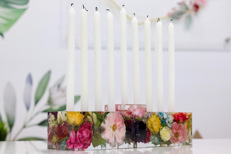 Op de afbeelding: Een bloemenkaarsenhouder van hars met negen witte kaarsen. De rechthoekige basis is gevuld met kleurrijke bloemen. De kaarsen zijn wit en zijn opgebrand. Een decoratief stuk voor de woondecoratie.