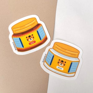 May include: Two sticker designs of a jar of tiger-themed peanut butter. The jar on the left is labeled "Red" and the jar on the right is labeled "White". Both jars have a blue and white label with the text "@riceclub.co" on the side.