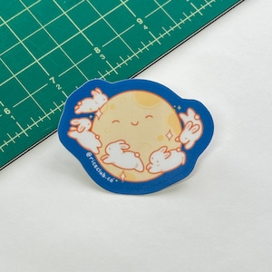 May include: A blue and yellow sticker with a smiling moon and five white bunnies surrounding it. The text "@riceclub.co" is at the bottom of the sticker.