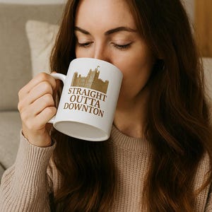 May include: White ceramic mug with a brown illustration of a castle and the text "STRAIGHT OUTTA DOWNTON". The mug has a curved handle and is being held by a person.