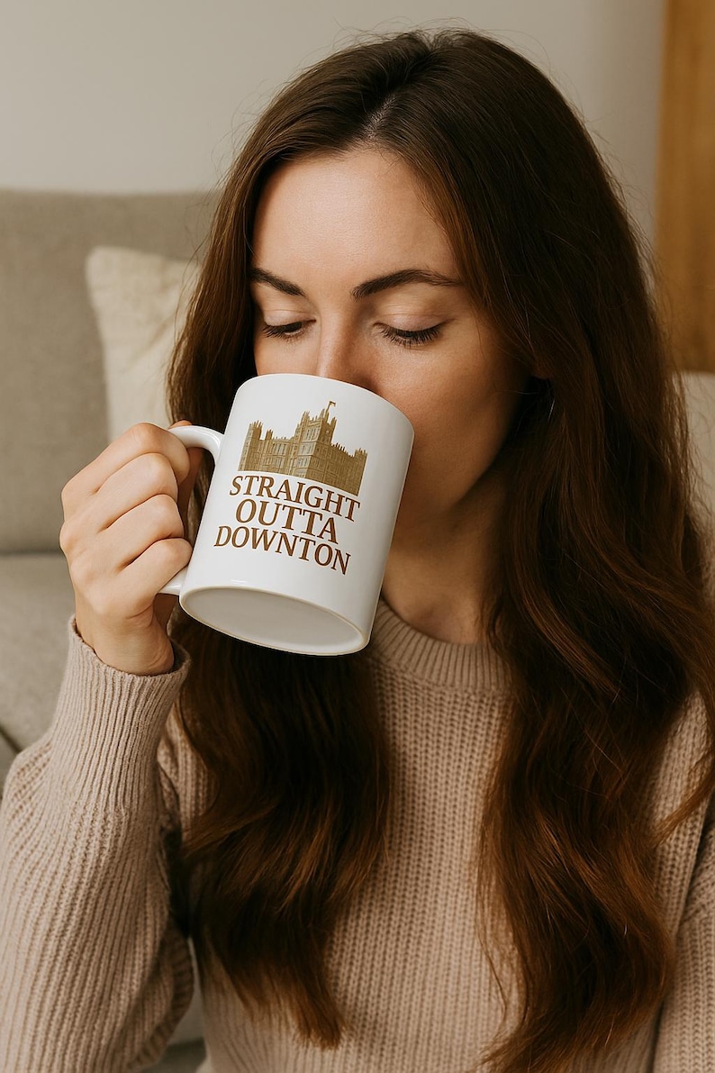 May include: White ceramic mug with a brown illustration of a castle and the text "STRAIGHT OUTTA DOWNTON". The mug has a curved handle and is being held by a person.