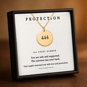 May include: A gold-toned circular pendant necklace featuring the number 444, displayed on a white card inside a black box. The card includes the text PROTECTION, 444 ANGEL NUMBER, and the message: You are safe and supported. The universe has your back.