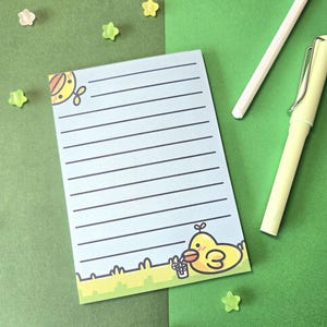 Könnte beinhalten: Ein hellblaues Notizbuch mit horizontalen Linien und einer Cartoon-Enten-Illustration. Das Notizbuch ist mit einer gelben Ente mit einem Korb verziert. Zwei Stifte und sternförmige Verzierungen sind ebenfalls zu sehen.