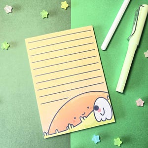 Könnte beinhalten: Ein gelber, liniertes Notizblock mit einer Cartoon-Hunde-Illustration in der unteren Ecke. Der Hund ist orange und weiß mit einem lächelnden Gesicht. Zwei Stifte und sternförmige Dekorationen sind ebenfalls zu sehen.