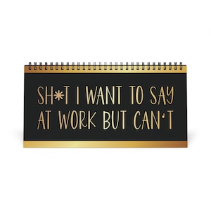 以下が含まれることがあります： 黒い背景と金の縁取りの卓上カレンダー。「SH*T I WANT TO SAY AT WORK BUT CAN'T」というテキストが金色のスクリプトで書かれています。カレンダーは上部がスパイラル綴じされています。