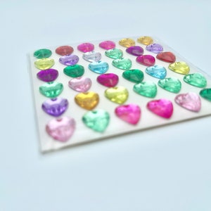 Op de afbeelding: Een vel met kleurrijke hartvormige stickers. De stickers zijn in rijen gerangschikt en zijn in verschillende tinten roze, rood, groen, blauw, geel en paars.