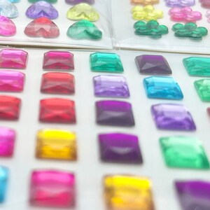 Op de afbeelding: Assortiment van kleurrijke, heldere en iriserende vierkante edelstenen. De edelstenen zijn gerangschikt in rijen op een witte achtergrond.