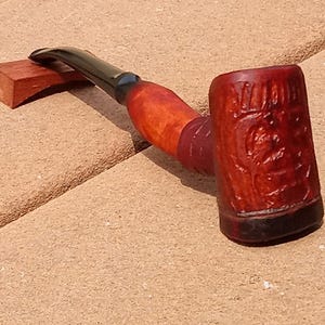 May include: A wooden smoking pipe with a black stem and a carved bowl. The bowl is decorated with a floral design and the words "William DeMuth" are carved into the side.