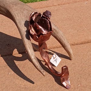 May include: A copper rose sculpture with a textured surface, mounted on a wooden antler. The sculpture is labeled "DA".