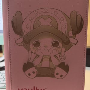 May include: A pink card binder featuring a graphic illustration of the character Tony Tony Chopper from One Piece on the front cover. The character is depicted with large eyes and antlers, wearing a hat. The brand name VaultX is printed in red at the bottom.