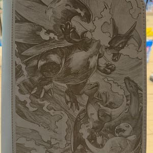 May include: A gray rectangular binder featuring a detailed laser-engraved illustration of three dragon-like creatures in various sizes. The artwork depicts a large winged creature soaring above two smaller creatures, all surrounded by stylized smoke and flames.