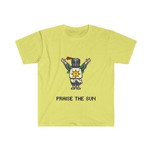 May include: Yellow t-shirt with a pixelated image of a knight in armor raising their arms and a sun with a smiley face on their chest. The text "PRAISE THE SUN" is printed below the image.