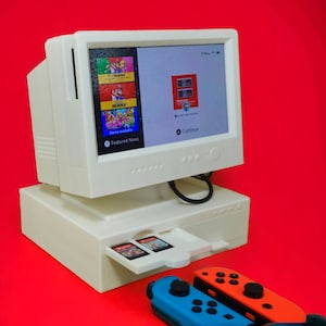 May include: A white desktop computer with a monitor displaying a menu with the words "Featured News" and "Continue". The computer has a slot for game cartridges and is sitting on a red surface. Two video game controllers are in the foreground.