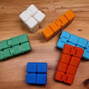 May include: Five fidget toys in different colours and designs. The toys are made of plastic and have a square shape. The colours are green, orange, white, blue, and red.