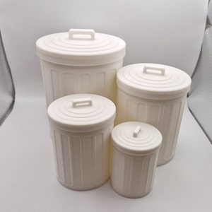 Peut inclure: Quatre mini-poubelles blanches de tailles différentes sont présentées. Chacune a un couvercle avec une poignée et des détails verticaux striés. La plus petite mesure environ 10 cm de haut.