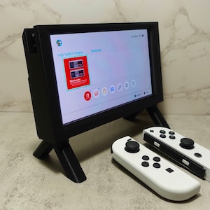 May include: A black Nintendo Switch console with a white screen displaying the Nintendo Switch Online menu. The console is sitting on a black stand with three legs. Two white Nintendo Switch Joy-Con controllers are lying on a white surface.