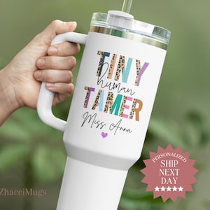 May include: White tumbler with a handle, featuring the text "TINY human TAMER Miss Anna" in colorful, playful lettering. The design includes leopard print accents and a purple heart. A straw extends from the lid.