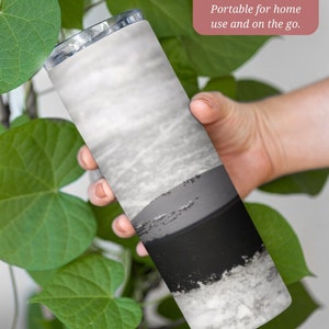 May include: A 20 ounce stainless steel tumbler with a black and white abstract design. The tumbler is held in a hand and is surrounded by green leaves. "20 oz. Size Tumbler. Portable for home use and on the go." is written in pink text on a white background.