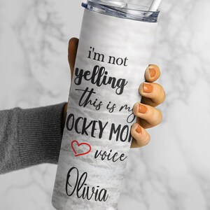 May include: White stainless steel tumbler with a marble pattern and black text that reads "I'm not yelling this is my hockey mom voice Olivia". A red heart is next to the word "voice".