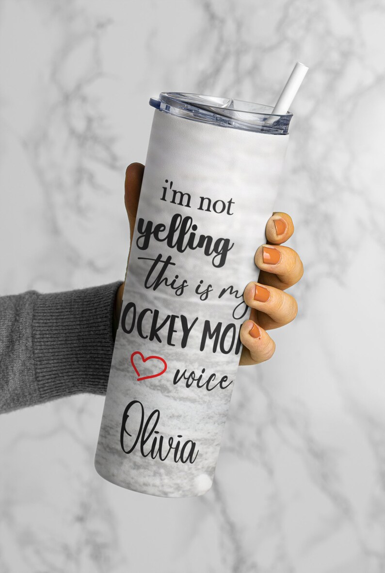 May include: White stainless steel tumbler with a marble pattern and black text that reads "I'm not yelling this is my hockey mom voice Olivia". A red heart is next to the word "voice".