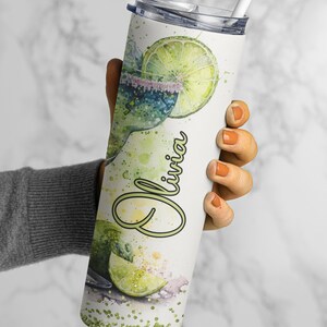 May include: A white stainless steel tumbler with a green and white watercolor design of a margarita with lime slices and the name "Olivia" written in green. The tumbler has a lid and a straw.