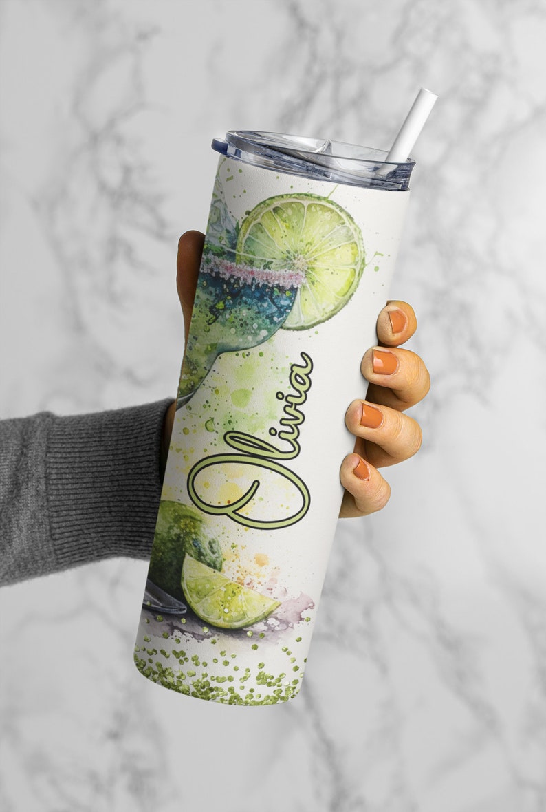 May include: A white stainless steel tumbler with a green and white watercolor design of a margarita with lime slices and the name "Olivia" written in green. The tumbler has a lid and a straw.