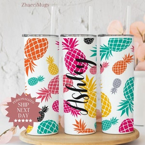 May include: Three stainless steel tumblers with a white background and a pineapple pattern in various colors. The tumblers have lids and straws. The middle tumbler has the name "Ashley" written on it in black cursive.