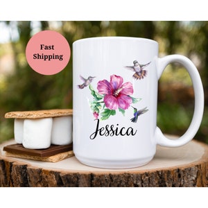 May include: White ceramic mug with a pink hibiscus flower and hummingbirds. The mug has the name "Jessica" written in black cursive.