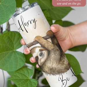 May include: A white 20 ounce stainless steel tumbler with a watercolor illustration of a sloth hanging from a branch. The tumbler has the text "Hurry" at the top and "Be" at the bottom.