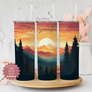 May include: Three stainless steel tumblers with a sunset mountain scene and the name "Samantha" on the middle tumbler. The tumblers are on a wooden surface.