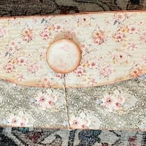 May include: A floral patterned envelope clutch with a round closure. The top flap features a cream background with pink flowers, while the bottom has a gray floral design. A string secures the closure. The clutch is a vintage-inspired accessory.