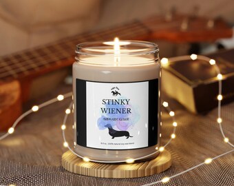 May include: A brown soy wax candle with a black label that reads "Stinky Wiener Aromatic Elixir" and features a watercolor illustration of a dachshund. The candle is on a wooden coaster and surrounded by white string lights.