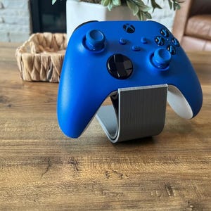 May include: A blue Xbox controller is displayed on a gray stand. The controller has a black faceplate and white accents.