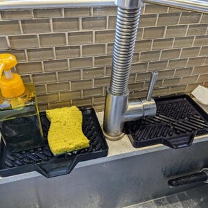 May include: A black dish drying rack with a yellow sponge and a yellow soap dispenser. The rack has a grid pattern and is designed to fit over a kitchen sink.
