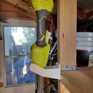 May include: A white plastic wall mount with a yellow and black Ryobi weed trimmer attached to it. The mount is attached to a wooden shelf.