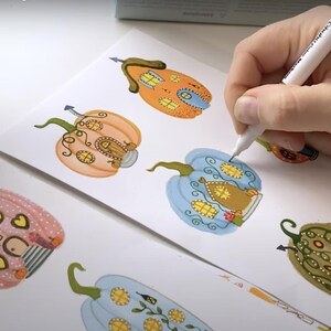 K&ouml;nnte beinhalten: Eine Hand zeichnet farbenfrohe K&uuml;rbisillustrationen mit einem wei&szlig;en Stift auf wei&szlig;em Papier. Die K&uuml;rbisse sind mit skurrilen hausartigen Details in Orange-, Rosa-, Blau- und Gr&uuml;nt&ouml;nen verziert. Im Hintergrund sind Marker zu sehen.