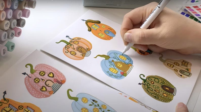 K&ouml;nnte beinhalten: Eine Hand zeichnet farbenfrohe K&uuml;rbisillustrationen mit einem wei&szlig;en Stift auf wei&szlig;em Papier. Die K&uuml;rbisse sind mit skurrilen hausartigen Details in Orange-, Rosa-, Blau- und Gr&uuml;nt&ouml;nen verziert. Im Hintergrund sind Marker zu sehen.