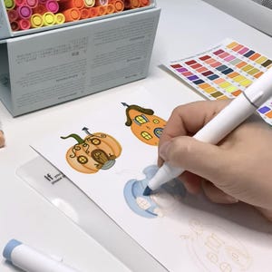 K&ouml;nnte beinhalten: Eine Person verwendet einen wei&szlig;en Marker, um eine Zeichnung eines K&uuml;rbis-Hauses auszumalen. Auf dem Tisch liegen mehrere wei&szlig;e Marker, zusammen mit einer Box mit Markern und Farbtafeln. Die Zeichnung befindet sich auf wei&szlig;em Papier.