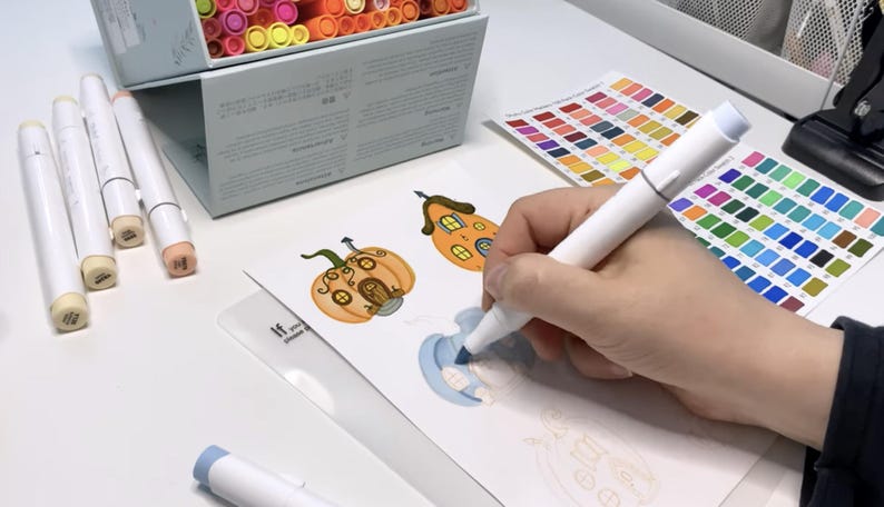 K&ouml;nnte beinhalten: Eine Person verwendet einen wei&szlig;en Marker, um eine Zeichnung eines K&uuml;rbis-Hauses auszumalen. Auf dem Tisch liegen mehrere wei&szlig;e Marker, zusammen mit einer Box mit Markern und Farbtafeln. Die Zeichnung befindet sich auf wei&szlig;em Papier.