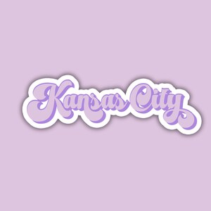 K&ouml;nnte beinhalten: Ein lilafarbener Aufkleber mit dem Text "Kansas City" in einer retro-kursiven Schrift.