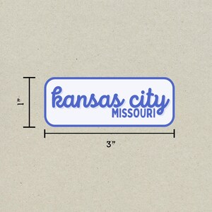 May include: A rectangular sticker with rounded corners, featuring the words "Kansas City MISSOURI" in blue script and block letters on a white background. The sticker measures 3 inches long and 1 inch high.