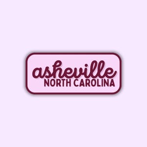 May include: A rectangular sticker with rounded corners, featuring the text "asheville NORTH CAROLINA" in a cursive font. The text is in a dark burgundy color, set against a light pink background, with a dark burgundy border.