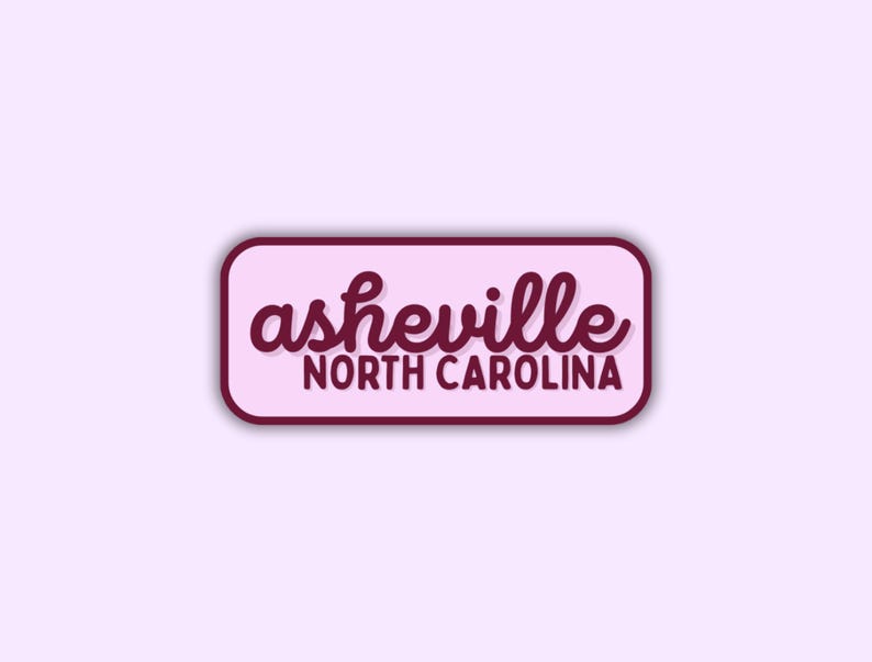 May include: A rectangular sticker with rounded corners, featuring the text "asheville NORTH CAROLINA" in a cursive font. The text is in a dark burgundy color, set against a light pink background, with a dark burgundy border.