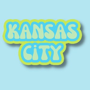 May include: A graphic design with the text "KANSAS CITY" in a retro, bubble-letter style. The letters are outlined in yellow and filled with light blue.