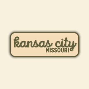 May include: A rectangular sticker with rounded corners features the words "Kansas City Missouri" in a cursive font. The text is in a dark olive green color, set against a beige background, with a dark green border.