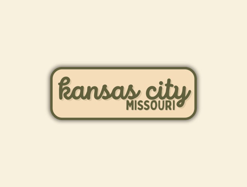 May include: A rectangular sticker with rounded corners features the words "Kansas City Missouri" in a cursive font. The text is in a dark olive green color, set against a beige background, with a dark green border.