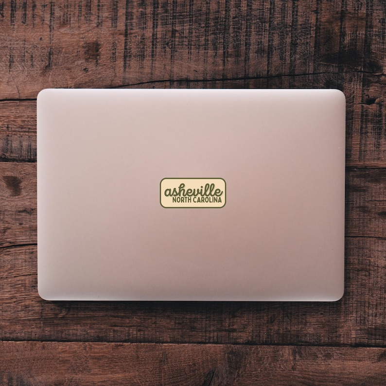 May include: A laptop with a sticker that reads "Asheville North Carolina" in a vintage style. The sticker is rectangular with a light yellow background and dark green text. The laptop is silver and sits on a wooden surface.