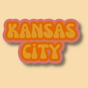 May include: A retro-style graphic design with the text "KANSAS CITY" in orange letters with a pink outline.