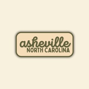 May include: A rectangular sticker with rounded corners. The sticker has a light tan background with a dark green border. The words "asheville" are in a cursive font, and "NORTH CAROLINA" is in a sans-serif font.