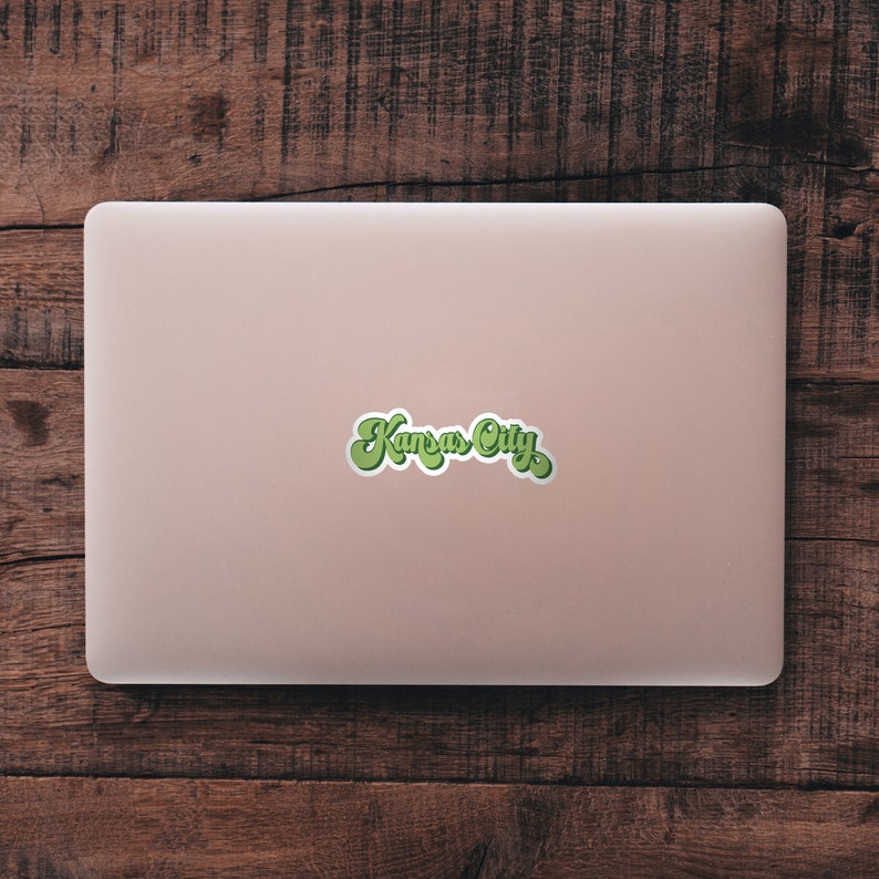 K&ouml;nnte beinhalten: Ein rosa Laptop mit einem gr&uuml;n-wei&szlig;en Aufkleber mit der Aufschrift "Kansas City" auf dem Deckel.